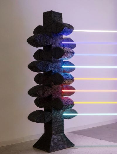 A textured black sculpture stands in a gallery, intersected by horizontal neon lights in blue, red, and yellow, creating a futuristic and dynamic feel.