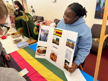 A young Black woman stands behind a table displaying a colorful poster board with pictures and text, and a rainbow-striped flag. She is looking at the poster board and holding up a piece of it.