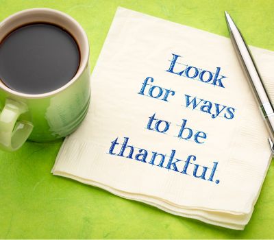 A cup of coffee near a napkin that reads "Look for ways to be thankful." with a pen nearby over a lime green background.
