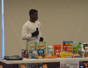 A young Black man in a white sweatshirt holds a microphone while standing behind a table of food products, including boxes of Chex and Ritz crackers, and various beverages. He appears to be giving a presentation or speech about the items.