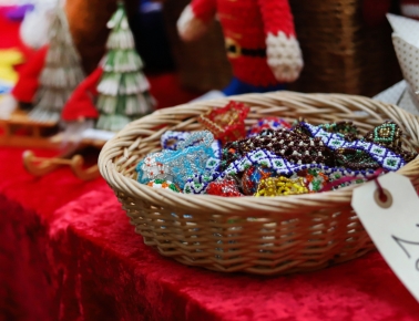 A woven basket overflowing with colorful, beaded Christmas ornaments, including stars and snowflakes, sits on a red velvet cloth table next to festive decorations.