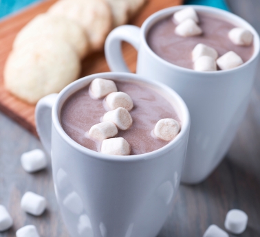 Two white mugs of steaming hot chocolate topped with marshmallows sit on a wooden surface, with a plate of cookies in the background.