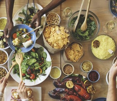 A diverse group enjoys a shared meal around a wooden table filled with colorful salads, chips, dips, and seafood, creating a lively and communal atmosphere.
