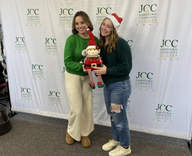 Two young women, one in a green sweater and light-colored pants holding an elf, and the other in a dark green sweater, jeans, and a Santa hat, pose in front of a backdrop with repeated "JCC Jamestown Community College" logos, likely at a holiday event.