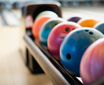 A row of colorful bowling balls rests on a rack at a bowling alley. Brightly lit, the scene conveys a vibrant and inviting atmosphere for a game.