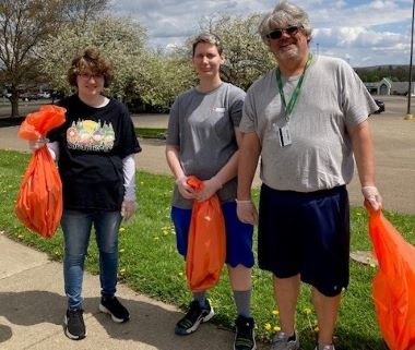 Three people stand outdoors holding orange trash bags, smiling for a photo. They're dressed casually, with blooming trees and a cloudy sky in the background.