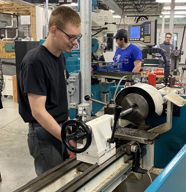 A man in glasses operates a metal lathe in a busy workshop. Two colleagues work on machines in the background, conveying a focused, industrious atmosphere.