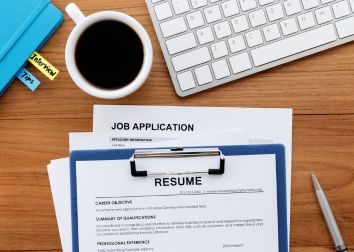 Overhead view of a paper resume, paper job application, blue folder, pen, mug filled with coffee, and keyboard sitting on a wood desk.