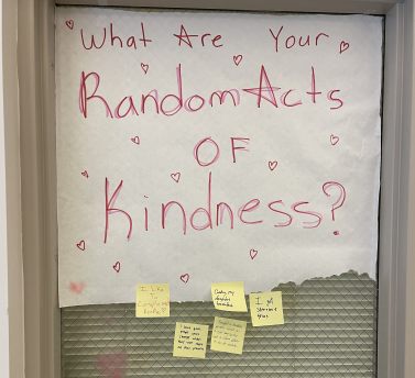 A whiteboard displays "What Are Your Random Acts of Kindness?" in pink marker. Several sticky notes with handwritten messages are attached to the bottom of the board.