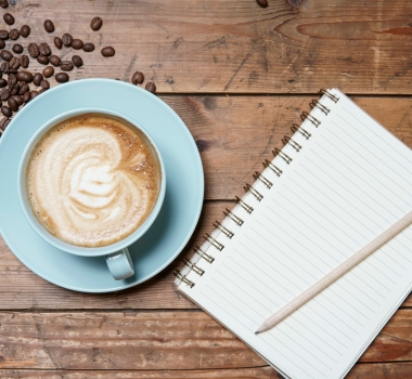 Overhead view of a latte with heart-shaped foam art, scattered coffee beans, and a pencil on a blank spiral notebook on a rustic wooden table, evoking a calm morning.
