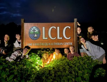 A group of smiling people pose around a "Lake Chautauqua Lutheran Center" sign at night, illuminated by light. The scene conveys joy and camaraderie.