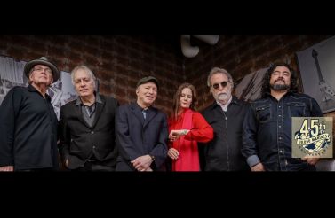 A diverse group of six musicians, including four men and two women, stand together against a brick wall. They are dressed in casual to semi-formal attire, with one woman in a bright red top. A circular emblem with "45th" and musical notes is visible on the right.