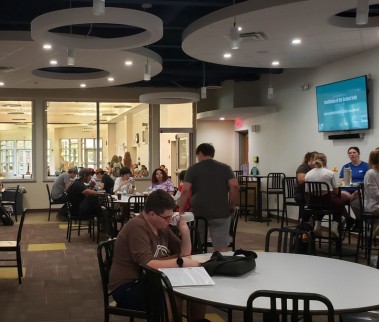 A bustling cafe or dining area with people seated at tables under modern circular ceiling lights. A screen displays information on the wall.