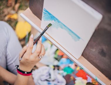 Close-up of a person's hands holding a paintbrush and applying light blue paint to a canvas on an easel, with a colorful palette of paints visible below.
