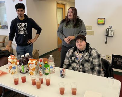 Three individuals stand and sit behind a table with various juice cartons and filled plastic cups. 