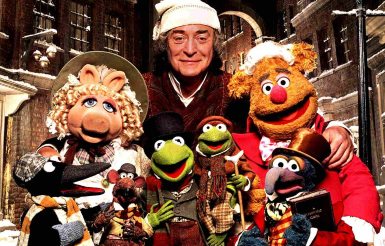 A group of Muppets and a man in a nightcap stand cheerfully on a snow-dusted street. They are dressed in festive, Victorian-style clothing.