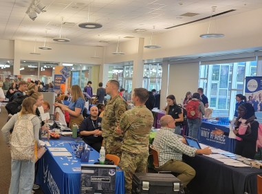 Military personnel and civilians gather at an indoor recruitment and information fair, talking at booths with blue tablecloths beneath bright ceiling lights.