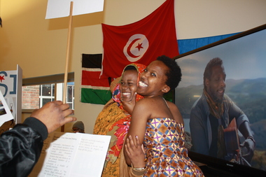 A couple of women in international clothing stand smiling under multiple international flags and in front of a large television screen that displays a man in an outdoor setting.