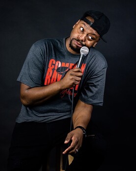 An image of comedian Ray Money holding a microphone on a dark stage or background, wearing a gray t-shirt with "Comedian Ray Money" printed in red vinyl, and a back baseball cap worn backwards on his head.