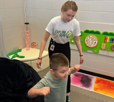 A young boy joyfully plays in a sensory room with a supervising adult. The room has colorful light panels, a maze toy on the wall, and a calming vibe.
