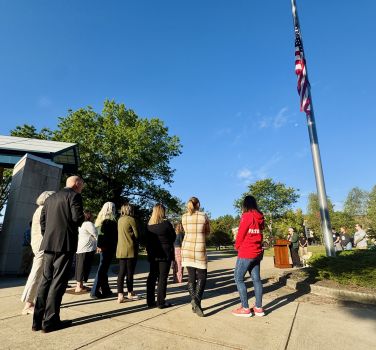 A group of people stand on a sidewalk, facing a flagpole with an American flag flying. The people are a mix of genders and ethnicities, and they are all dressed in casual attire. The flag is a symbol of patriotism and freedom.