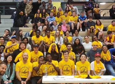 A large group of young adults in athletic apparel pose for a photo in a gymnasium. They are wearing yellow t-shirts with a hawk mascot.