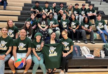 Group of students and adults sit on bleachers, wearing matching green shirts with a mascot. They appear cheerful and are posing with a banner.