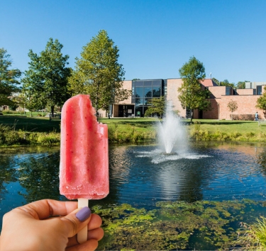 A hand holding a red popsicle near a pond on a college campus.