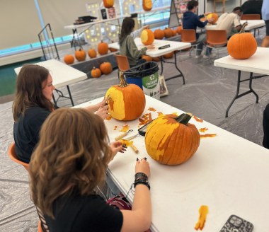Students carve pumpkins at white tables in a bright classroom, with carving tools and orange gourds nearby, creating a lively, creative atmosphere.
