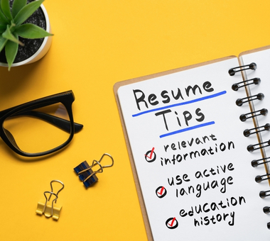 A notebook titled “Resume Tips” lists relevant information, active language, and education history; glasses, binder clips, and a plant sit on a bright yellow desk.