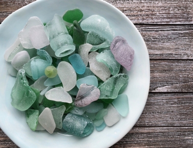 A light blue ceramic bowl overflows with a collection of colorful, frosted sea glass in various shades of green, blue, purple, and clear.
