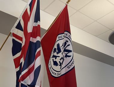 Two flags hang indoors: one is the British flag with red, white, and blue colors; the other is red, representing the Seneca Nation, with a circular emblem featuring text.
