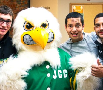 A cheerful group photo featuring three smiling young men posing with a large JCC Jayhawk mascot in a green "JCC" jersey. One man gives a thumbs up.
