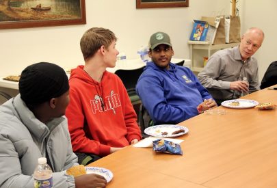 Four people sit at a table chatting over food. They appear relaxed and engaged, with plates and snacks in front of them. 