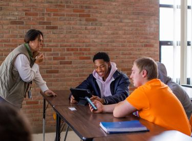 A teacher and three students gather around a table, looking at a tablet. The students are engaged in a lesson with the teacher.