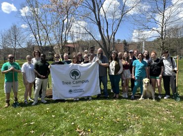 A group of people stands on grass holding a "Tree Campus" banner under a clear sky. Trees and a building are in the background; a dog sits near them.