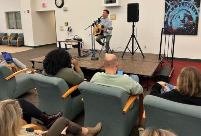 A singer performs on a small stage with an acoustic guitar, facing a relaxed audience seated in cushy chairs. A colorful abstract painting is on the wall.
