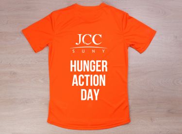 An orange t-shirt with "JCC SUNY HUNGER ACTION DAY" printed in white text on the back. The t-shirt is laid flat on a wooden surface.