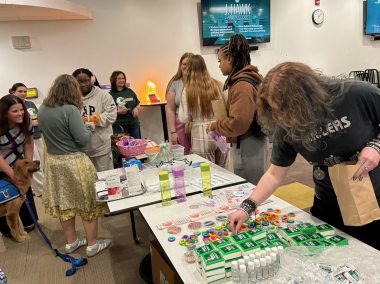 A diverse group of people stands around tables filled with various products and promotional materials, including small bottles, buttons, and green packaged items, at an indoor event.