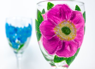 A wine glass painted with a pinkish purple flower with another glass painted with a blue flower in the background.