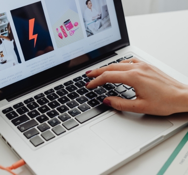 A hand types on a silver laptop displaying a website with product and lifestyle images, suggesting focused online browsing or shopping in a bright workspace.