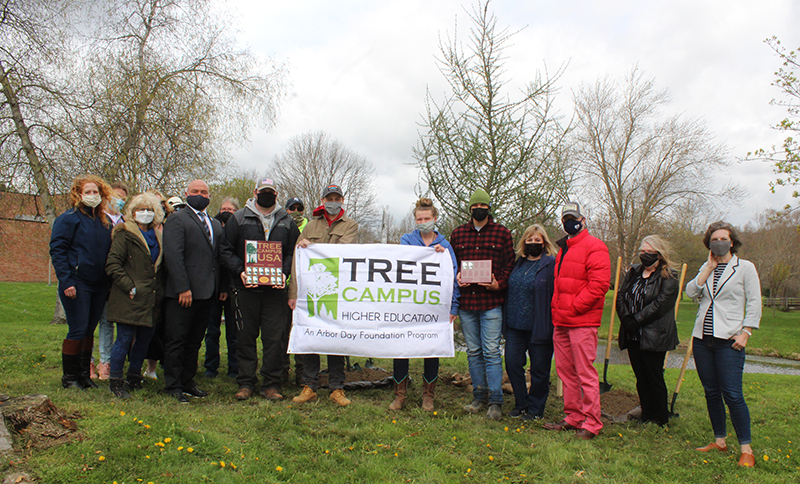Students, employees, and community members stand around a tree with a Tree Campus Higher Education sign.