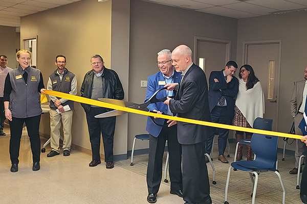 Two men cut a yellow ribbon with big scissors in front of onlookers at a grand opening ceremony.