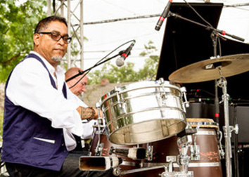 A man sitting at a set of drums with a microphone in front of him looks at the camera.