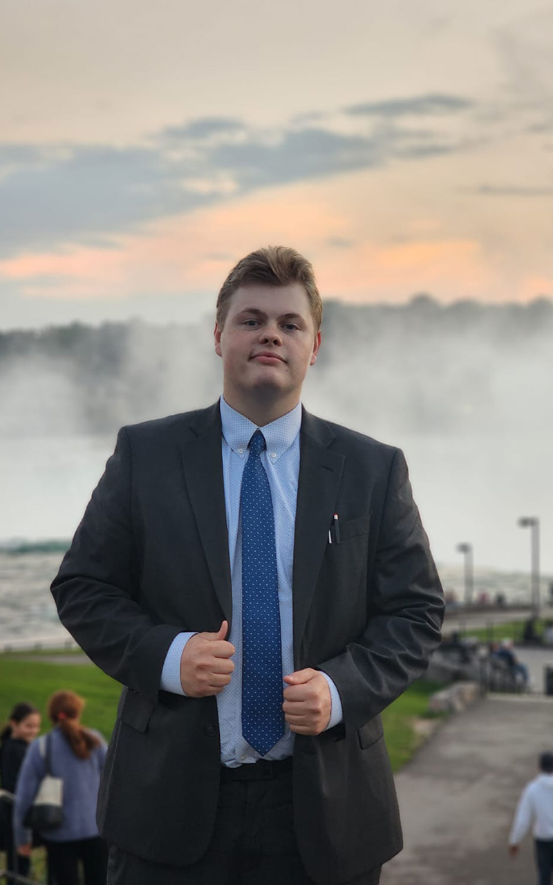 A man stands in front of Niagara Falls gently holding both lapels of his suit jacket.