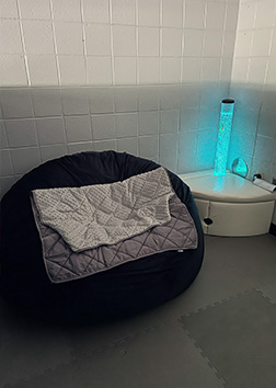 A dimly-lit area with corner table, glowing lava lamp, bean-bag chair and weighted blanket.