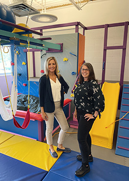 Two women stand in front of an indoor play area that utilizes a climbing wall, slide and swings for occupational therapy for children.