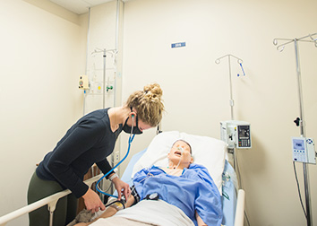 A nursing student is wearing a mask and leaning over a robotic patient, taking its pulse.