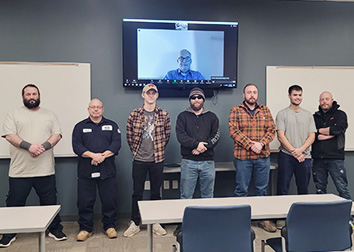 A group of students stand under a flat-screen monitor with a man pictured and there are tables and chairs in rows in the foreground.