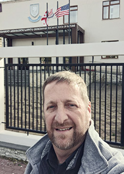 A man taking a selfie in front of a metal gate with a building with flags in the background.
